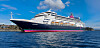 Et stort cruiseskip ligger til kai ved en kysthavn med blått hav og klar himmel.