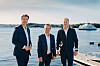 Tre menn står på en brygge med vann og en ferge i bakgrunnen.