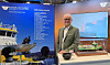 Mann står ved messestand med modellskip, skjermer og informasjonsplakater.