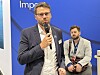 Mann med mikrofon taler foran en blå konferansevegg, mens en annen mann sitter bak ham.